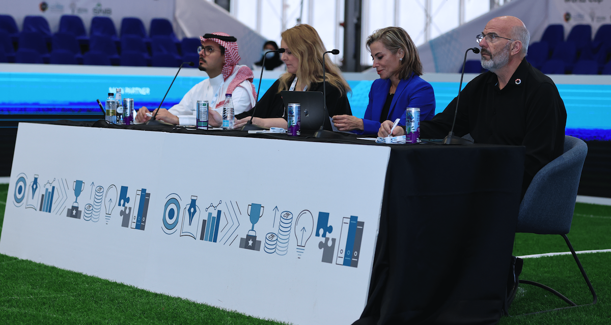 Panel of judges including middle eastern man in traditional garb, blonde woman in black jacket, brunette woman in blue jacket, and white man in black jacket.