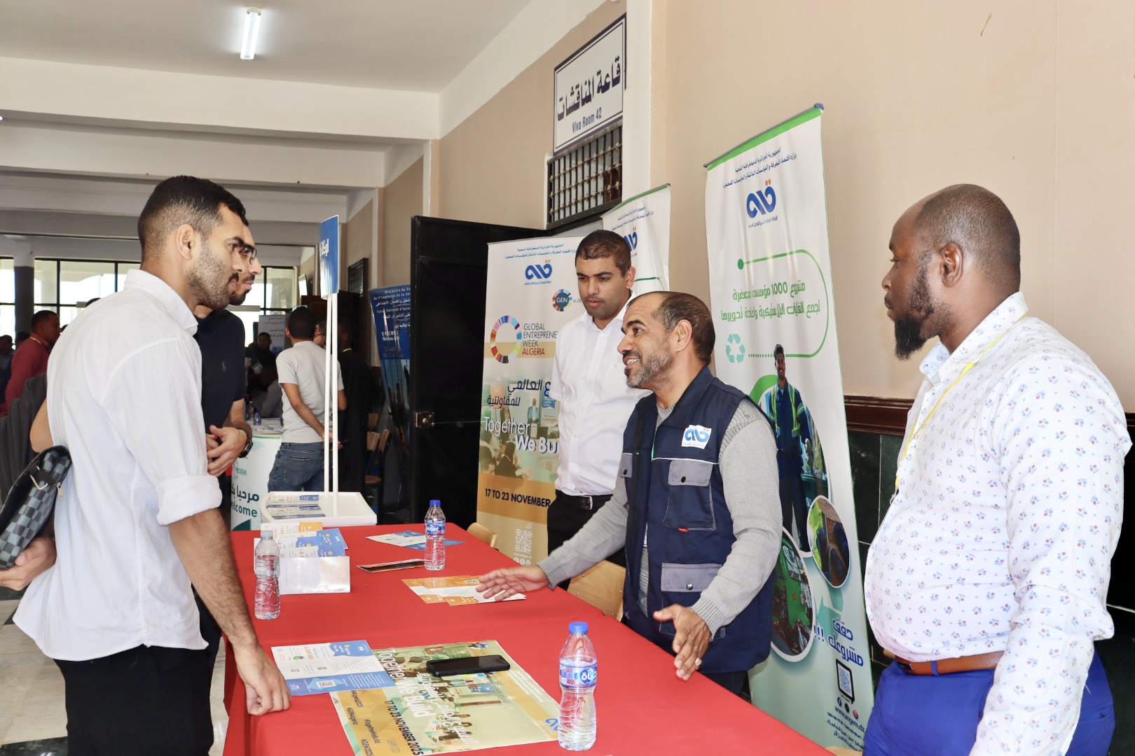 Image of a student approaching the booth of the National Agency for Microcredit Management (ANGEM) during the professional exhibition, listening to explanations provided by the institution’s representative about support and financing mechanisms.